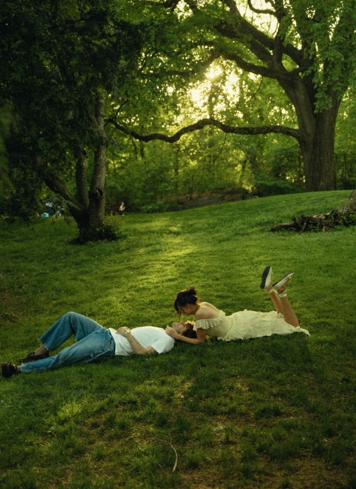 two people laying on the ground reading books in a park with green grass and trees