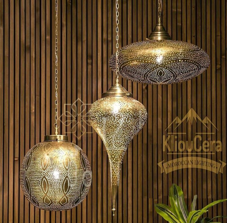 three gold colored lamps hanging from chains next to a potted plant and wooden wall