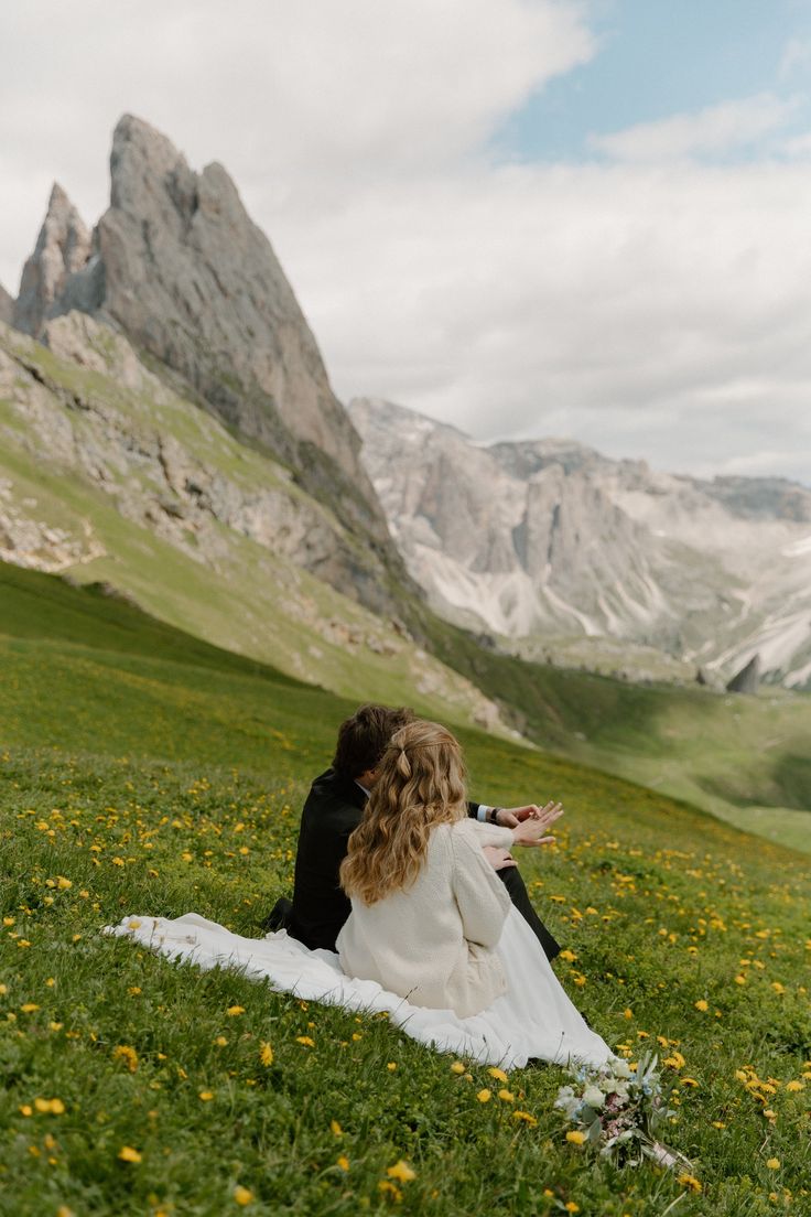 a bride and groom sitting in the grass looking at each other with mountains in the background