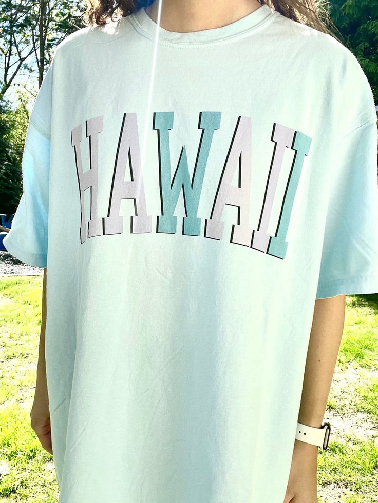 "Such a cute tee for summer! Comfort Colors chambray (light blue) t-shirt with \"HAWAII\" in blue and purple, with a black outline. Model is 5'4\" and wearing a size XL for an oversized look. 100% cotton." Light Blue Short Sleeve T-shirt For Summer, Casual Light Blue T-shirt For Summer, Blue Slogan T-shirt For Summer, Light Blue T-shirt With Letter Print For Spring, Trendy Blue Pre-shrunk T-shirt, Blue Graphic Tee With Slogan, Blue Graphic Tee T-shirt With Slogan, Graphic Tee Short Sleeve T-shirt, Light Wash Short Sleeve Graphic Tee