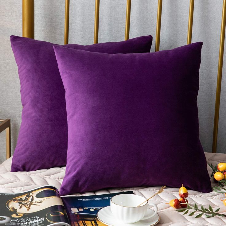 two purple pillows sitting on top of a bed next to a cup and saucer