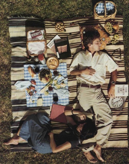 two people laying on a blanket in the grass with food and drinks laid out around them