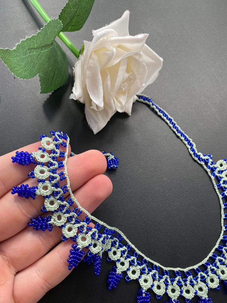 a hand holding a blue and white beaded necklace next to a flower on a table