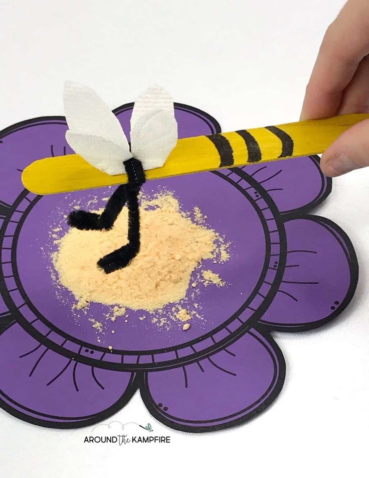 a hand is holding a pencil over a paper flower with a bee on it and powdered sugar in the middle