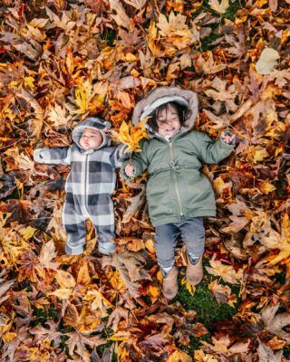 two young children laying in leaves on the ground with their faces covered by autumn leaves