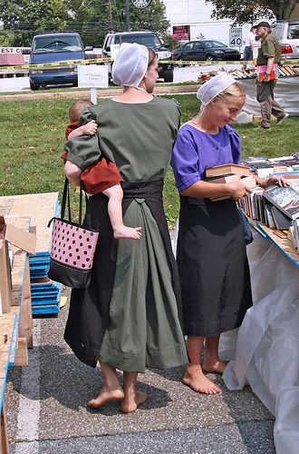 two women in dresses are looking at books on a table with other people standing around