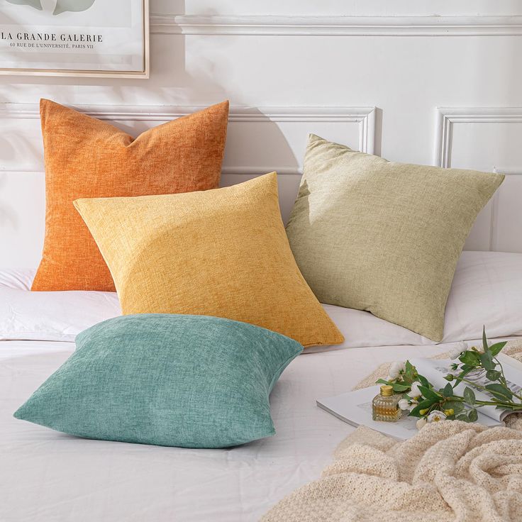 four different colored pillows sitting on top of a white bed next to a vase with flowers