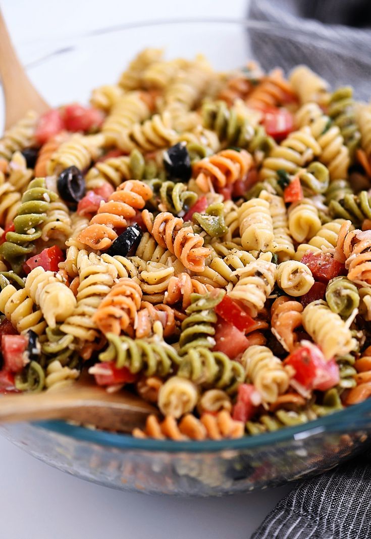 a glass bowl filled with pasta salad on top of a table next to a wooden spoon