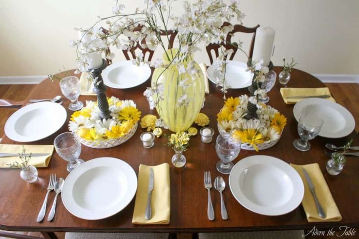 the table is set with yellow and white dishes, silverware, and flowers in vases