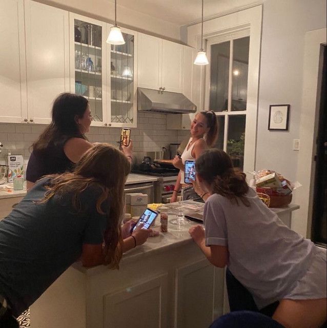 three women are standing at the kitchen counter and looking at their cell phones in front of them