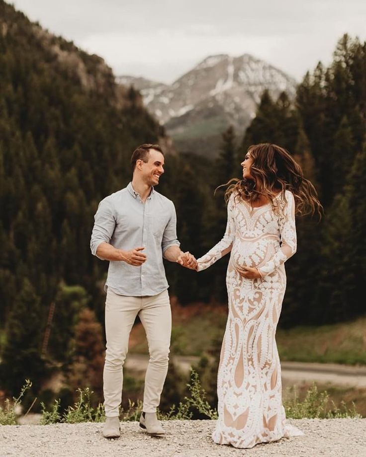 a man and woman holding hands while standing in front of trees with mountains behind them