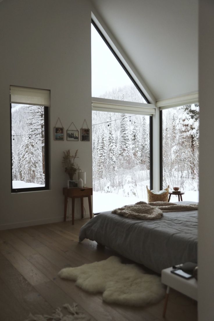 a bedroom with large windows and a bed in the middle, surrounded by snow covered trees