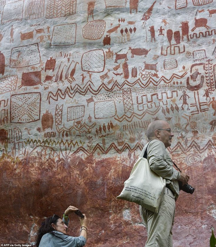 two people standing in front of a rock wall with paintings on it and one person holding a camera