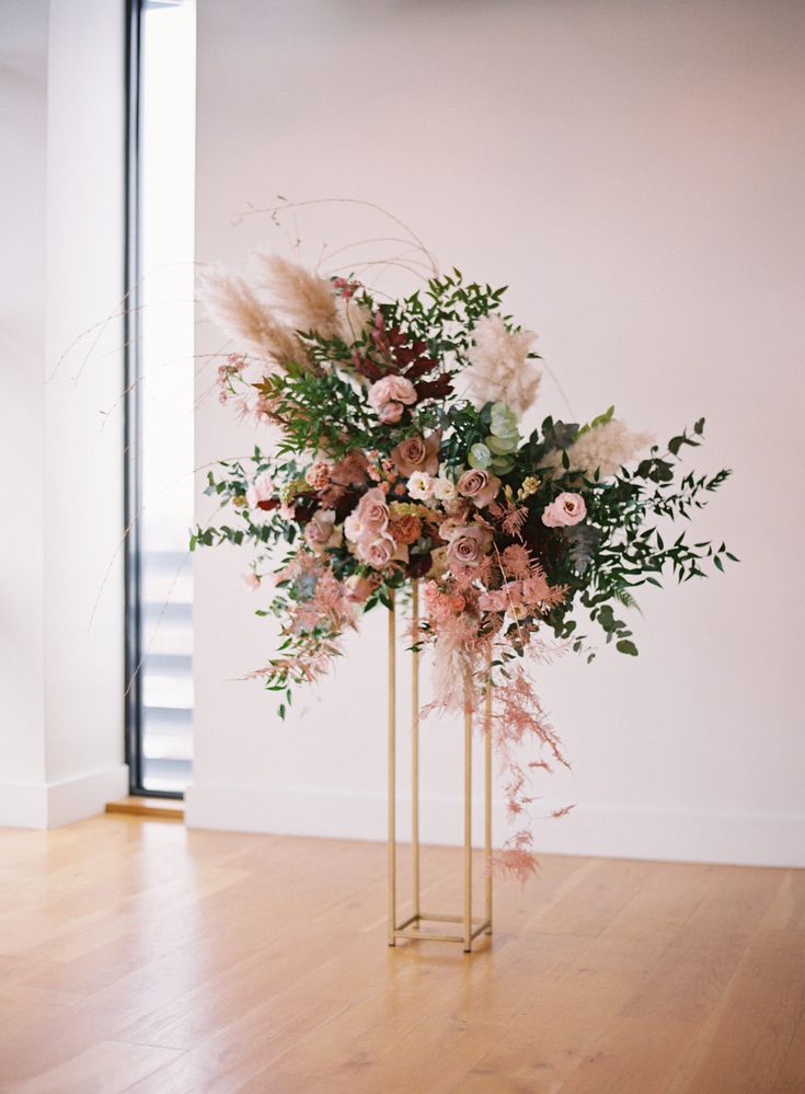 a tall gold vase with pink flowers and greenery in it on a wooden floor