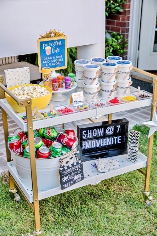 an outdoor movie party with popcorn, soda cans and snacks on a cart in the grass