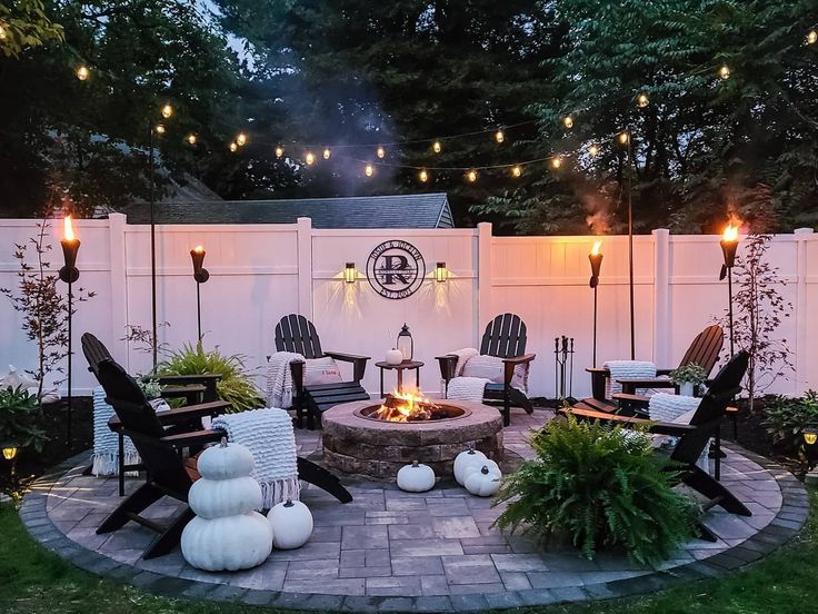 an outdoor fire pit surrounded by lawn chairs and string lights with pumpkins around it