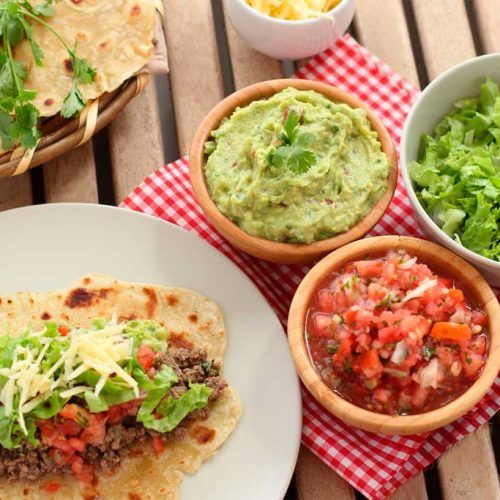 some tacos and salsa are sitting on a table with bowls of salad, tortilla chips and guacamole