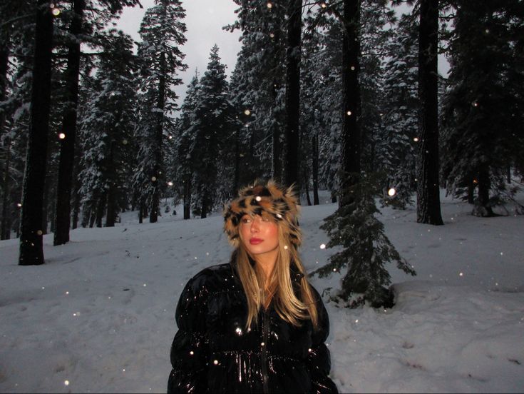 a woman standing in the snow wearing a fur hat and black coat with trees behind her