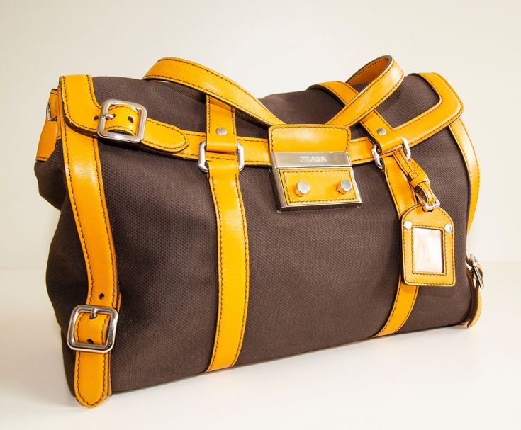 "An authentic Prada shoulder bag. The bag features a brown canvas exterior, orange leather trim, and silver-tone hardware. The interior is lined with yellow leather and there is one zipped pocket. The interior is neat & clean. The exterior is in very good condition, the trim has minor signs of use on the bottom and corners of the bag. Overall the bag is in really good condition and comes with its Prada cards and dust bag. Dimensions (cm & inch) Width: 40 cm / 15,7\" Height: 23,5 cm / 9,3\" Depth Brown Satchel With Silver-tone Hardware For Travel, Brown Coated Canvas Shoulder Bag With Palladium Hardware, Orange Travel Bag With Silver-tone Hardware, Cognac Travel Bag With Silver-tone Hardware, Yellow Coated Canvas Shoulder Bag For Travel, Brown Travel Bags With Silver-tone Hardware, Travel Bags With Palladium Hardware In Brown, Yellow Travel Shoulder Bag With Palladium Hardware, Designer Orange Satchel For Travel