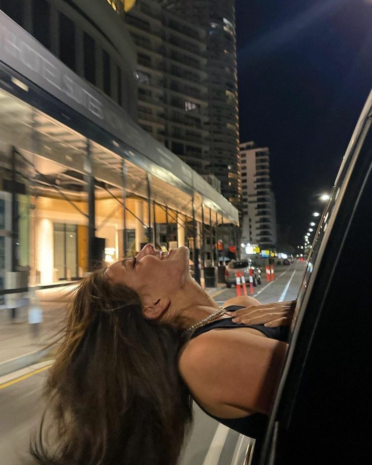 a woman leaning out the window of a car on a city street at night with buildings in the background