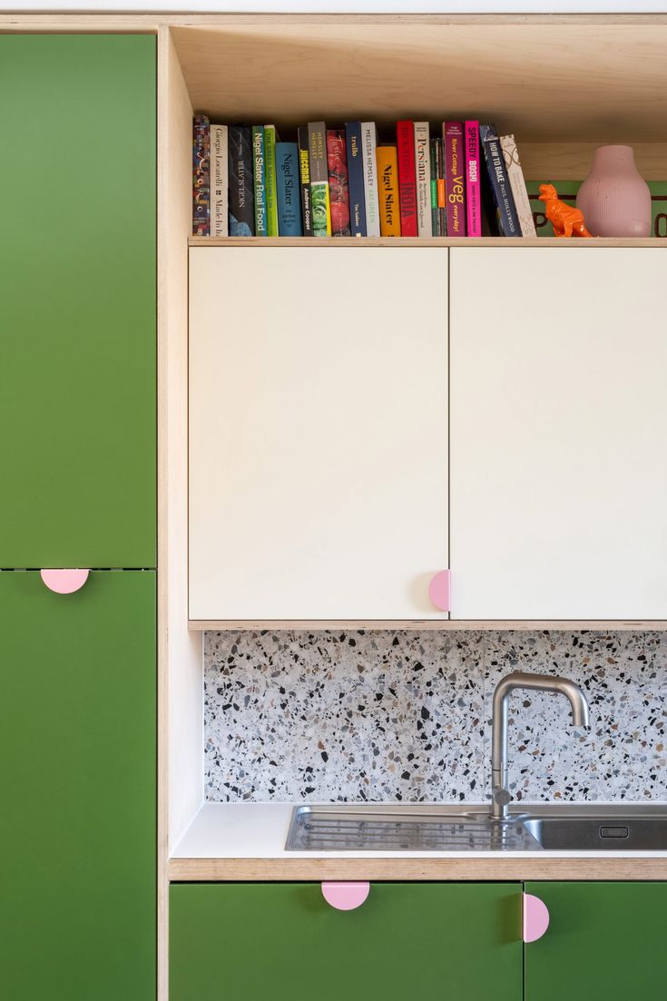 a kitchen with green cabinets and white cupboards next to a stainless steel sink in front of a bookshelf