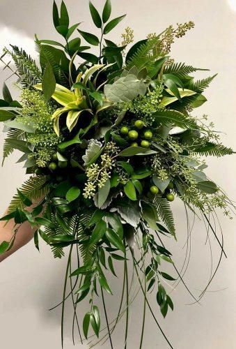 a bouquet of green leaves and greenery on a white surface with a hand holding it