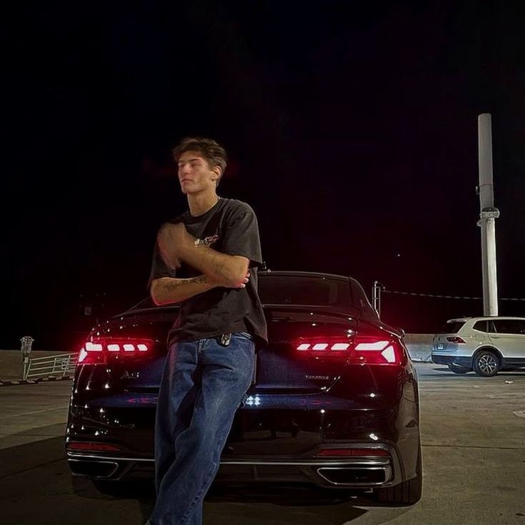 a man leaning on the hood of a black car in a parking lot at night