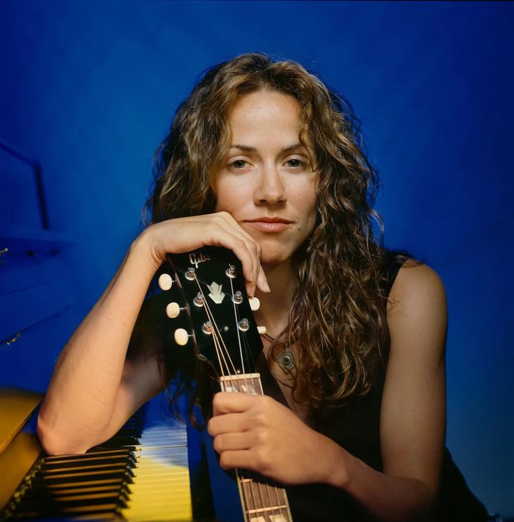 a woman with long hair is holding an acoustic guitar and posing for a photo in front of a piano