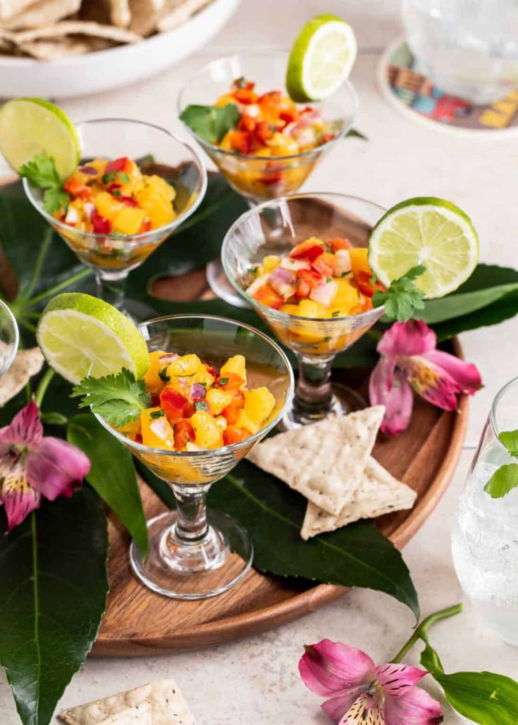 small glasses filled with fruit and garnish on a wooden tray next to flowers