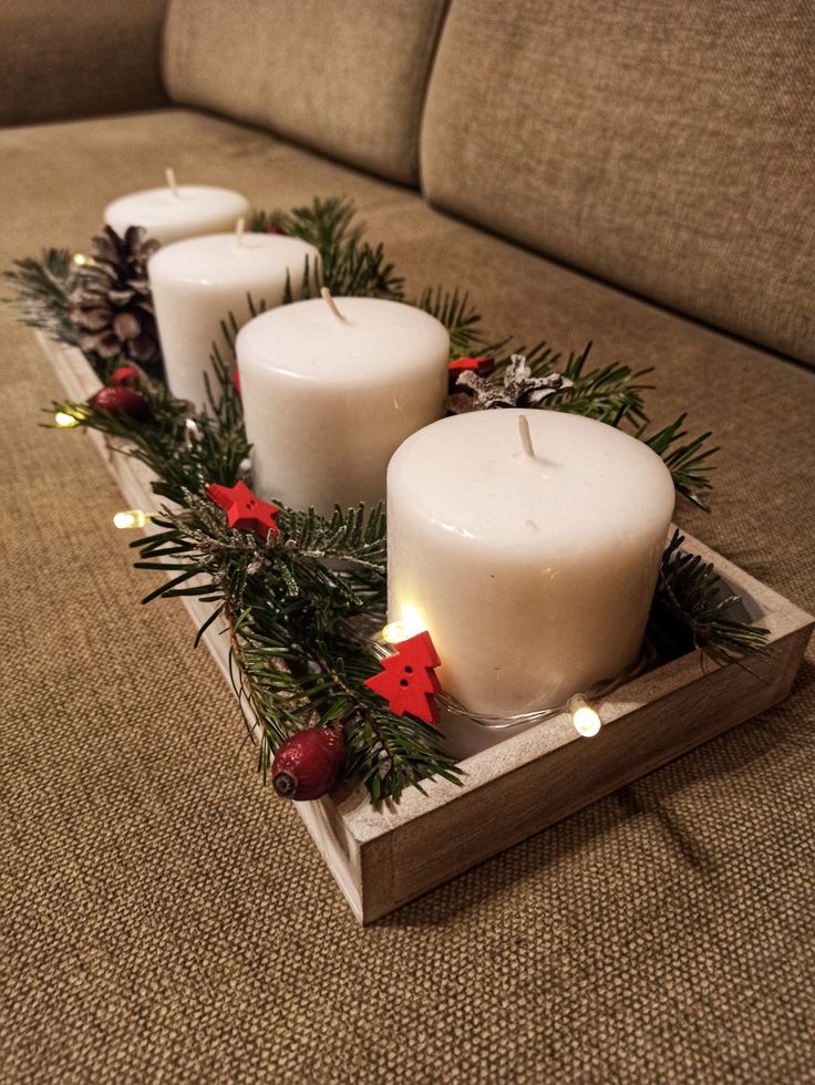 three white candles sitting on top of a wooden tray filled with greenery and pine cones