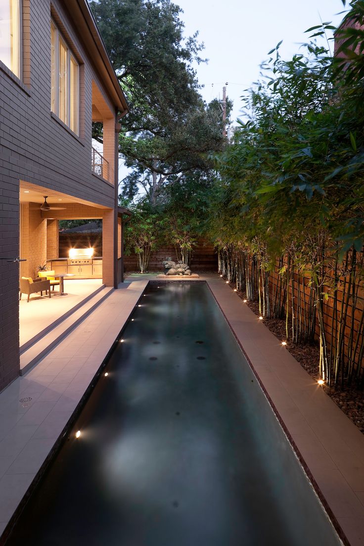 an outdoor swimming pool surrounded by bamboo trees and lights in the evening time, next to a house