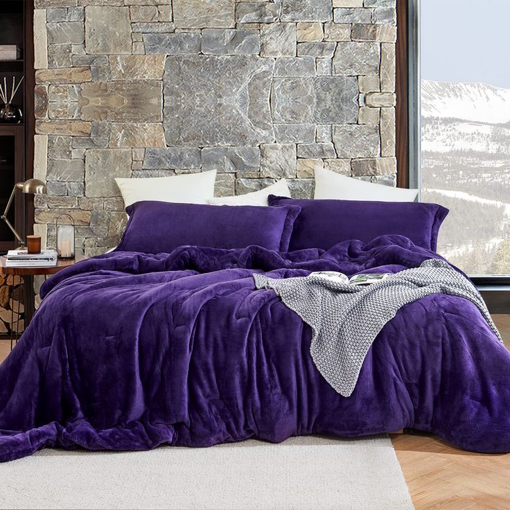 a bed with purple comforter and pillows in front of a stone wall