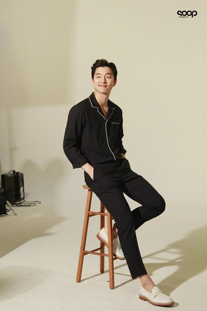 a man sitting on top of a wooden stool in front of a white wall wearing black pajamas