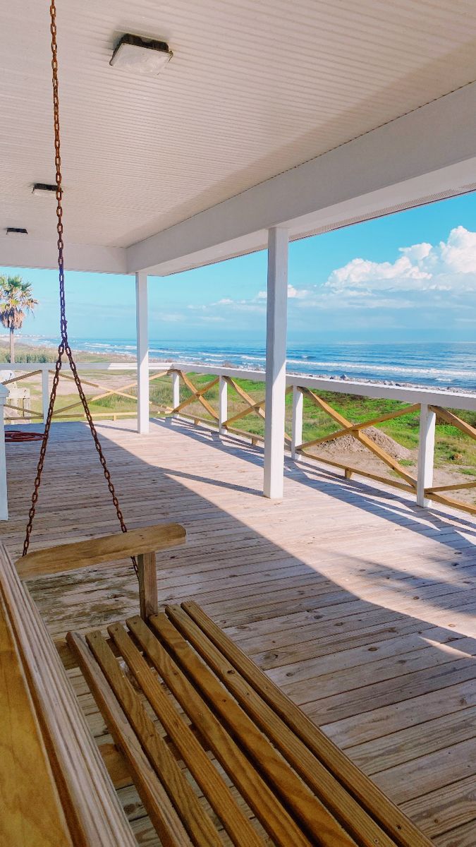 a wooden porch with a swing hanging from it's side and the ocean in the background