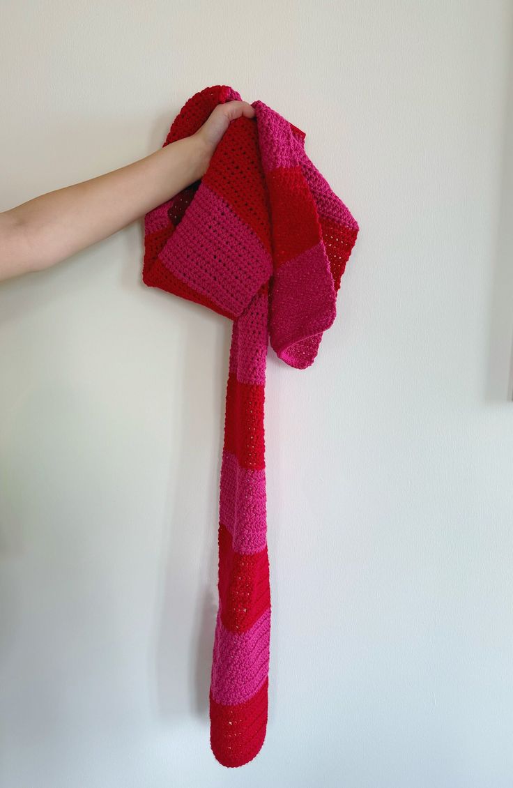 a person is holding a pink and red knitted scarf on the wall with their hand