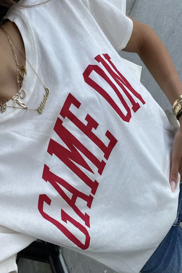 Get ready to level up your wardrobe with the Game On Boyfriend Tee! This stylish white tee featuring bold red writing and a fun "Game On" graphic will have you looking and feeling confident. Whether you're hitting the gym, running errands, or just lounging around, this tee is the perfect blend of comfort and style. White Varsity T-shirt With Screen Print, Athleisure Graphic Print T-shirt For Game Day, Trendy White T-shirt With Lettering, White Logo Print T-shirt For Game Day, Trendy Red T-shirt For Sports, Trendy Letter Print Workout T-shirt, White Text Print T-shirt For Game Day, White Graphic Print Muscle Tee For Streetwear, White Cotton Muscle Tee With Graphic Print