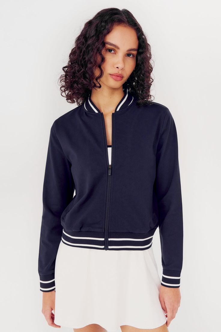 Woman wearing an Ever Supplex Jacket in Indigo by SPLITS59 with white trim and a white spandex skirt. Stretch Track Jacket With Ribbed Cuffs For Sports, Sporty Long Sleeve Outerwear With Ribbed Waistband, Sporty Track Jacket With Ribbed Cuffs For Loungewear, Sporty Long Sleeve Activewear With Elastic Cuffs, Relaxed Fit Athleisure Track Jacket With Ribbed Cuffs, Sporty Relaxed Fit Track Jacket For Loungewear, Sporty Relaxed Fit Outerwear With Ribbed Collar, Sporty Outerwear With Ribbed Collar And Relaxed Fit, Sporty Outerwear With Baseball Collar For Spring