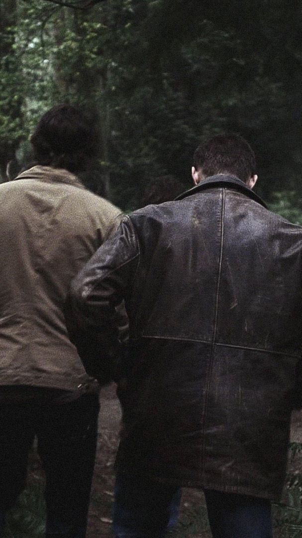 two men walking in the woods with one holding his hand out to another man who is wearing a brown leather jacket