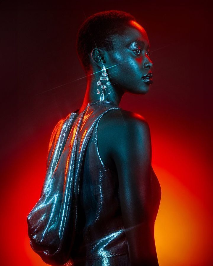 a woman with black skin wearing shiny clothing and large earrings, standing in front of a red background