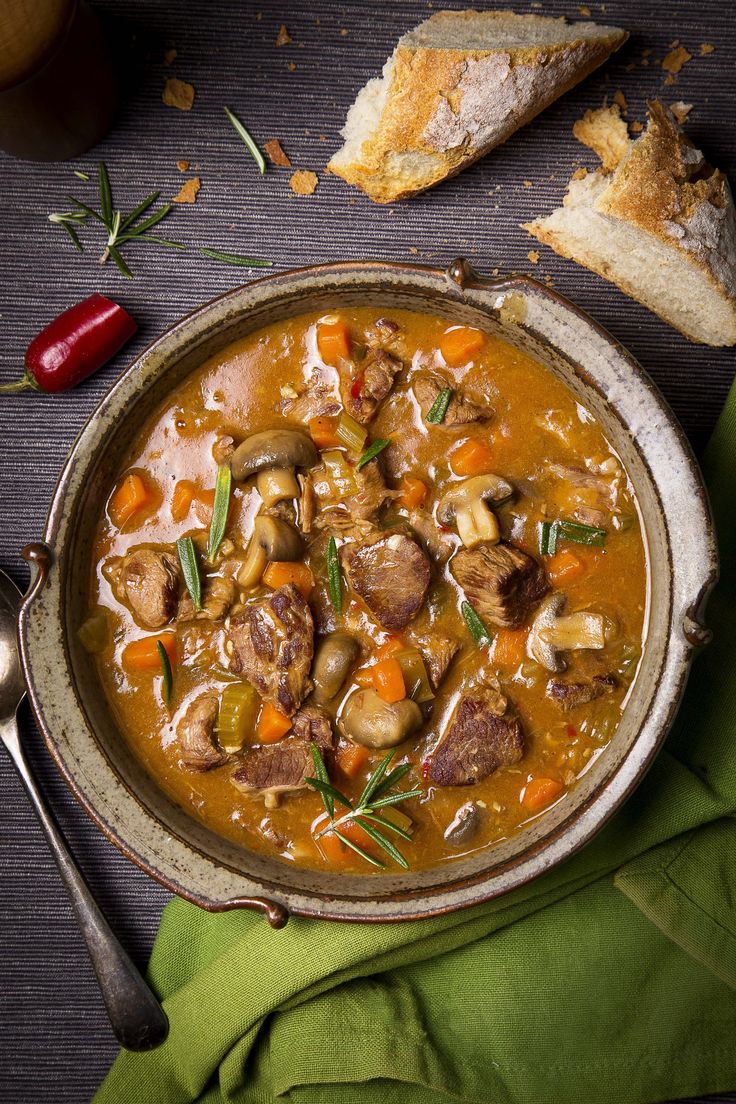 a bowl of soup with meat, vegetables and bread on a table next to a spoon