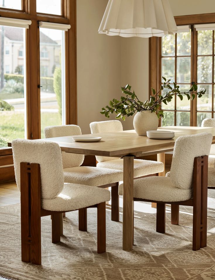 a dining room table with chairs and a vase on top of it in front of large windows