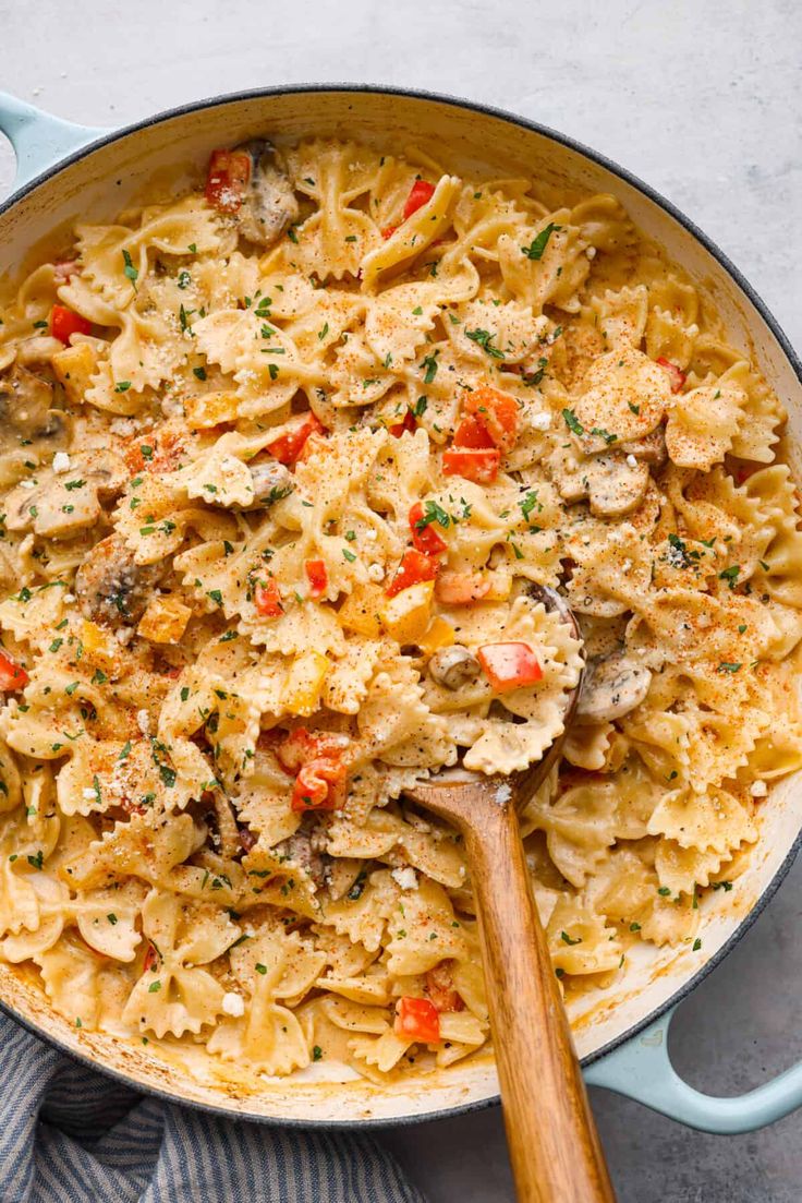 a skillet filled with pasta and vegetables on top of a blue towel next to a wooden spoon