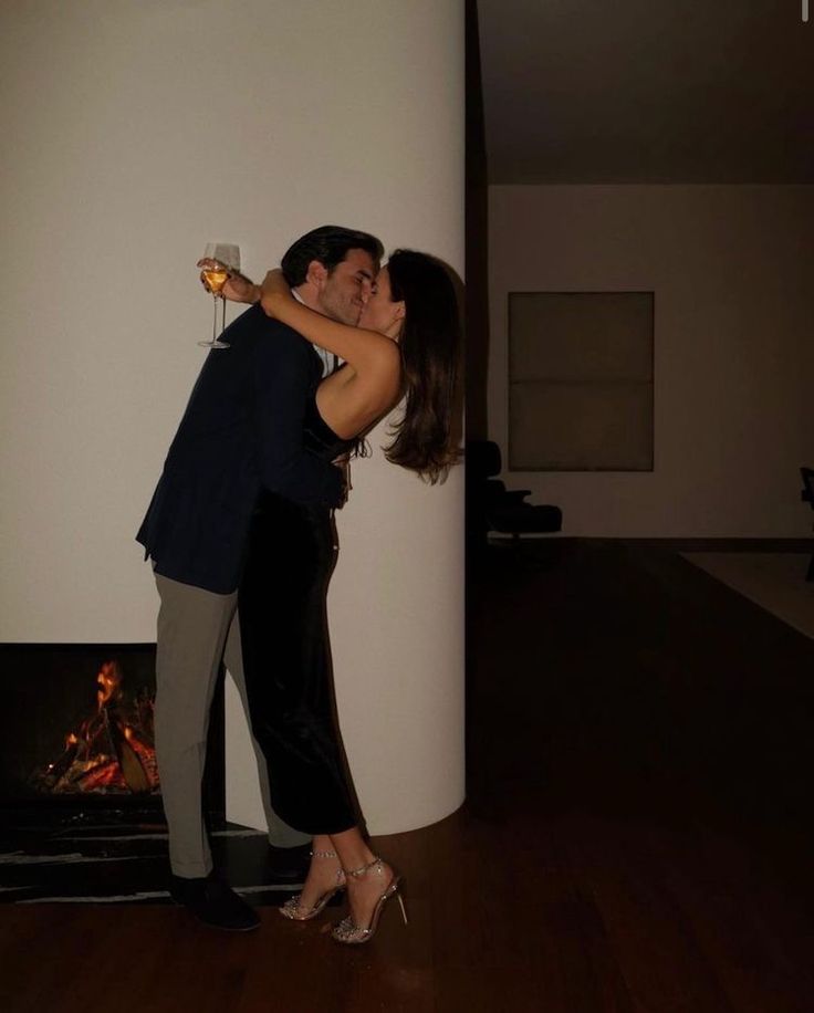 a man and woman kissing in front of a fire place with their arms around each other