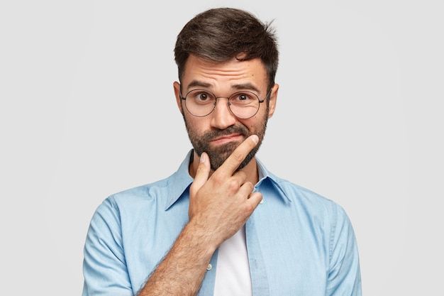 a man wearing glasses and a blue shirt is holding his hand to his chin while looking at the camera