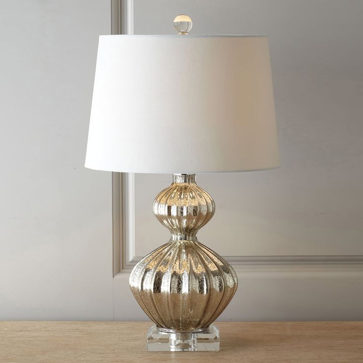 a lamp on a table in front of a window with a glass base and white lampshade