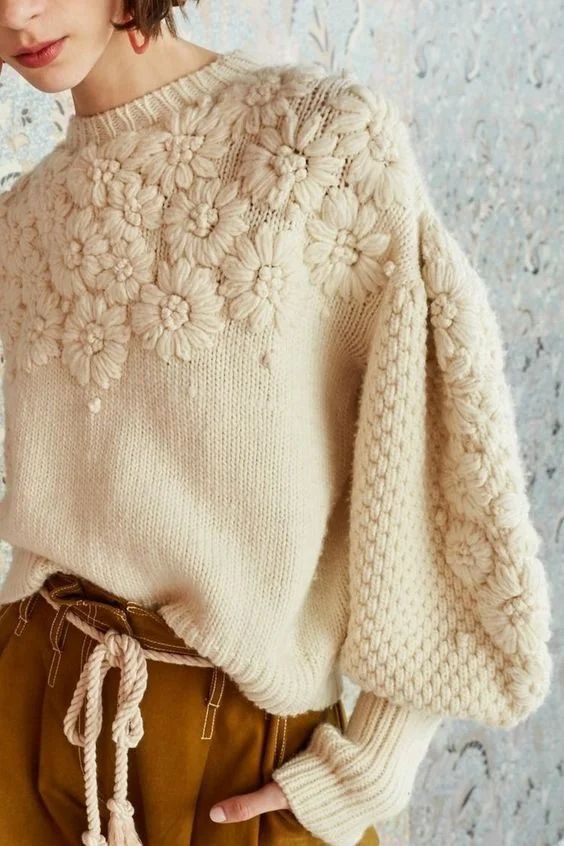 a woman is standing in front of a wall wearing a white sweater and brown pants