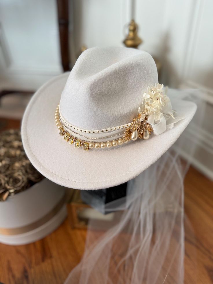 Handmade custom white bridal cowboy hat decorated with beautiful pearly and bling hat bands, rhinestones, letters, and preserved flowers.  This hat can be created with a distressed look, but will closely resemble the images provided. While some cards, ribbons, feathers, and other embellishments may differ slightly, the customizations will remain very similar to those depicted in the photos. Each hat can be shipped in a custom hat box that protects hat shape and features while shipping to you. The hat box is perfect to store your hat in when not worn. Should you choose to ship in a hat box, please add the item to your cart as well. (Price is not included with the purchase of this hat.) If you would like to request additional features or modifications, please do not hesitate to message AD yo Cowboy Hat Decorated, Cowboy Hat With Veil, Bride Cowboy Hat, Embellished Cowboy Hat, Cowboy Hut, Bling Hat, Veiled Hats, Hat Bands, Chapeau Cowboy