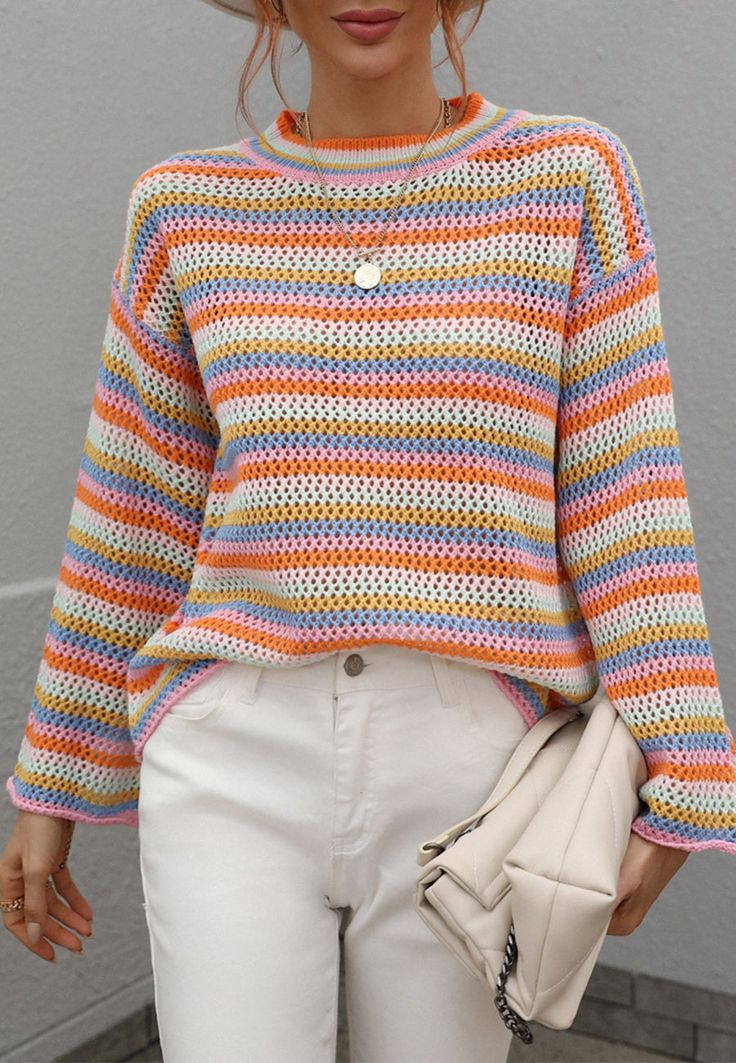 a woman wearing white pants and a multicolored knitted sweater is standing in front of a gray wall