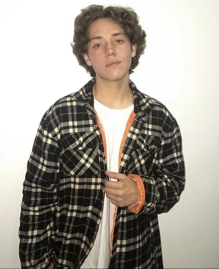 a young man standing in front of a white wall wearing an orange and black checkered shirt