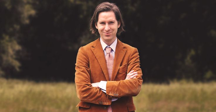 a man with his arms crossed wearing a brown suit and tie, standing in front of a grassy field